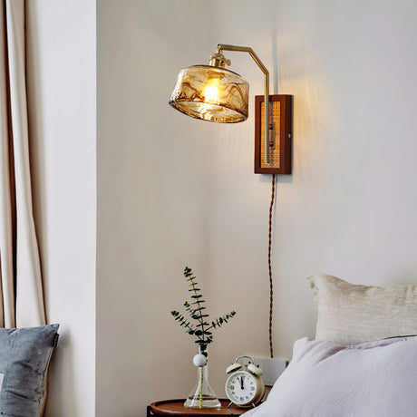 The sconce with an amber drum shade above a nightstand with a clock beside a bed