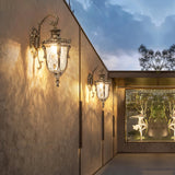 The lantern lit on a wall under a timber pergola beside a gate at dusk
