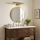 Brass bar lit above a round mirror over a walnut vanity with towels and a plant