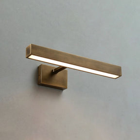 The shortest brass bar lit on a pale wall, the block bracket under its middle