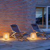 Lanterns lit on paving beside deck chairs against a brick house wall