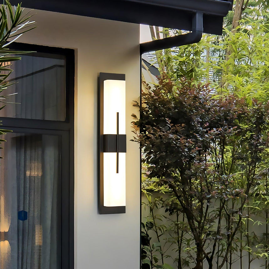 The bar light on a white wall beside a glass door under an eave, shrubs to the side