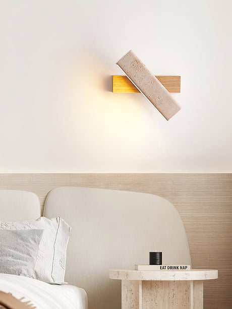 Sconce lit above a bed with pale cushions and a round stone side table