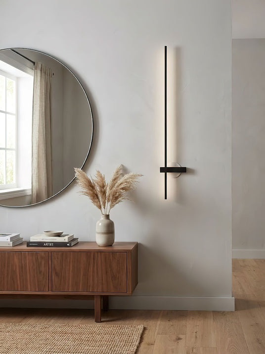 Lit beside a round mirror above a timber sideboard with a vase of grasses
