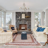 Hung in a room with a stone fireplace, armchairs, a patterned rug and windows
