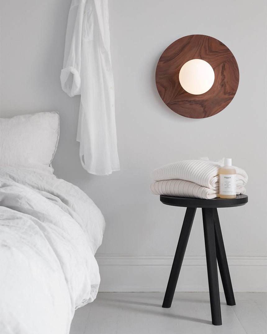 Lamp on a bedroom wall above a black stool with a jar and a folded towel
