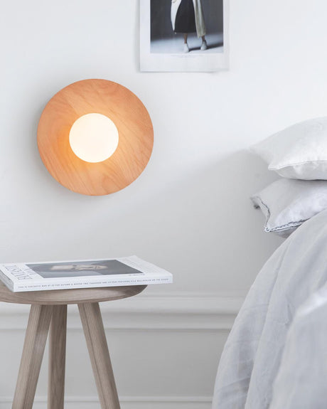 Lamp lit above a bed beside a wood stool holding a book and pale bedding