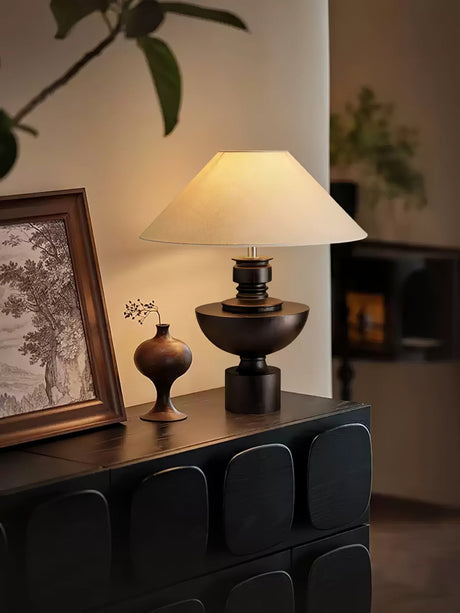 Beige table lamp lit on a sideboard beside a small dark vase