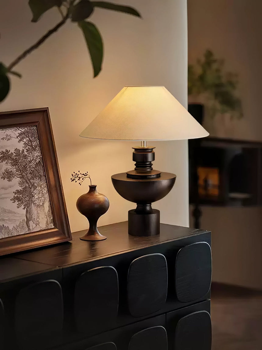 Beige table lamp lit on a sideboard beside a small dark vase