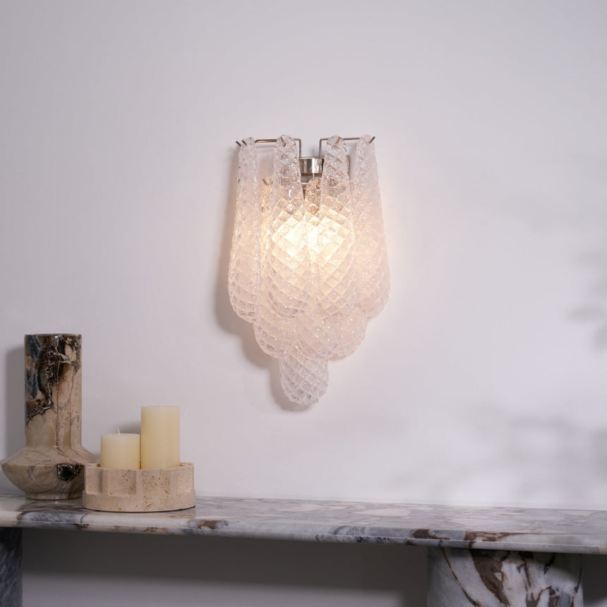 The clear sconce lit above a stone shelf with candles and a vase