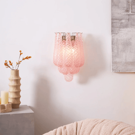 The pink sconce lit above a side table with a ribbed vase and dried branches