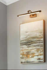 Bronze bar lit above a tall framed canvas on a pale wall seen from the side