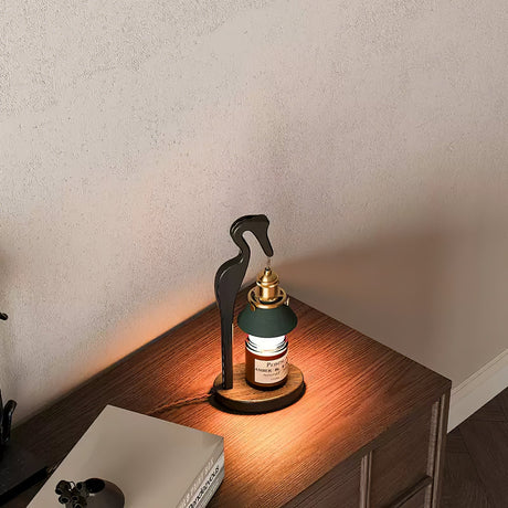 Heron lamp lit on a desk beside a laptop and a pale wall