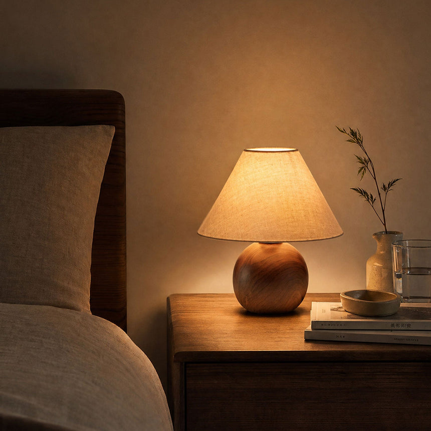 Ceramic bedside lamp lit on a nightstand beside pillows and a sprig