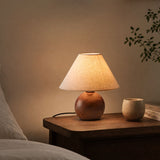 Ceramic bedside lamp lit on a wooden table beside a bed and a cup