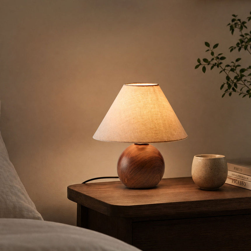 Ceramic bedside lamp lit on a wooden table beside a bed and a cup