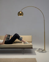 Brass arc lamp reaching over a low daybed, a reader lying under the dome