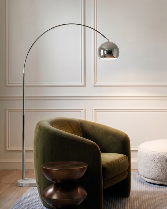 Chrome arc lamp leaning over a green velvet armchair beside a paneled wall