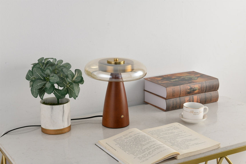 Clear glass table lamp lit in cool white beside books and a plant