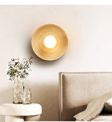 Sconce lit above a round side table with a vase of leaves beside a bed