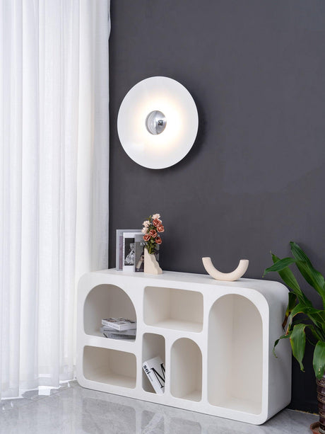 The white disc lamp on a dark wall above a white shelf with a plant
