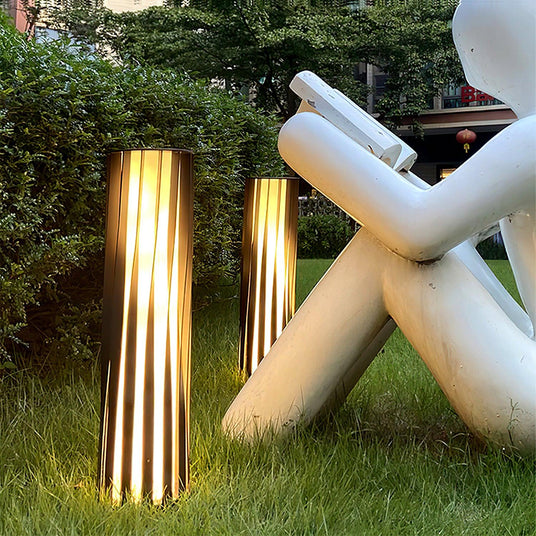 Two lit on a lawn beside a pale sculpture with a hedge behind them