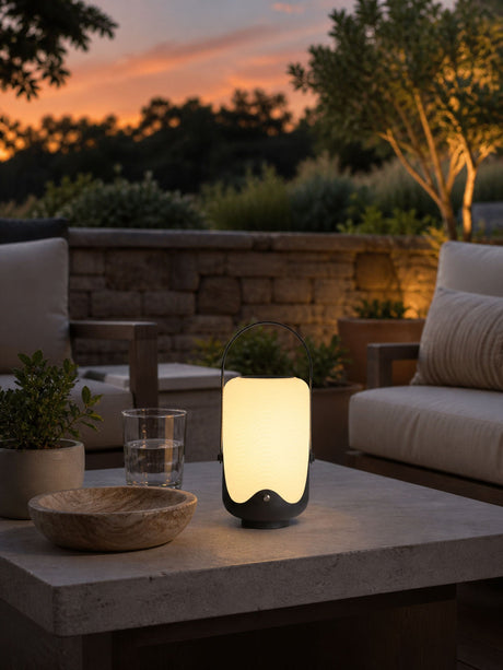 The lamp lit on a stone terrace table with a garden wall behind at dusk