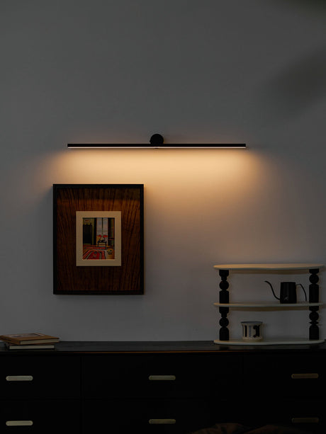 Black bar lit above a framed print on a dark wall, warm light washing down it