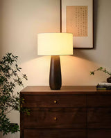 Cream table lamp lit on a dark dresser beside a green plant