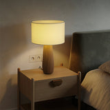 Cream table lamp lit on a wood nightstand beside a made bed
