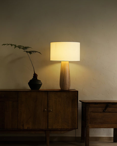 Cream table lamp lit on a sideboard beside a small dark vase