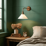 The sconce lit on a green paneled wall above a nightstand with a clock and dried flowers