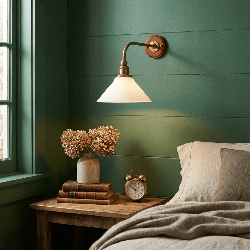 The sconce lit on a green paneled wall above a nightstand with a clock and dried flowers