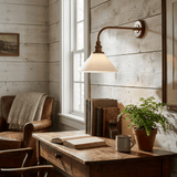 The sconce lit above a wood desk with an open book and a plant beside a window