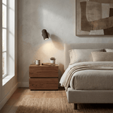 Sconce lit beside a bed above a pale wood nightstand with a cup and book