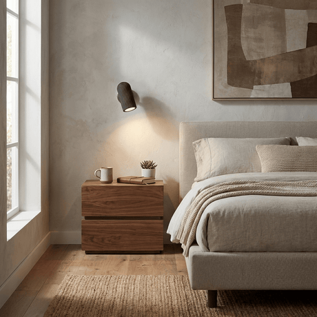 Sconce lit beside a bed above a pale wood nightstand with a cup and book