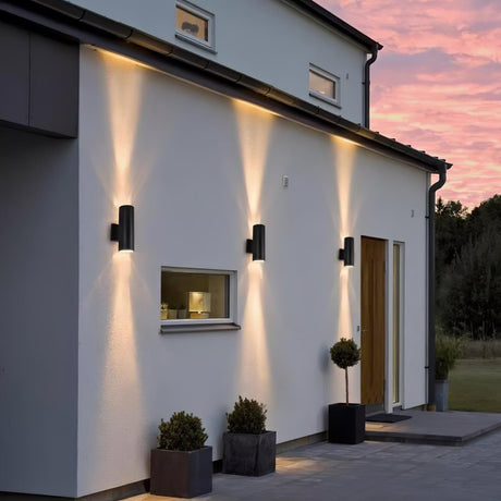 A row of cylinder lights along a white facade at sunset, planters below