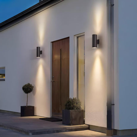 Two cylinder lights on a white facade on either side of a wood front door with potted trees