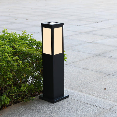 The bollard unlit on a pale paved walk beside a clipped green shrub