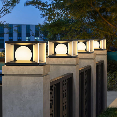 Lit cubes on pillars beside a white fence and a planted driveway edge