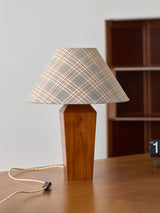 Plaid shade lamp unlit on a wood desk in front of a dark bookcase