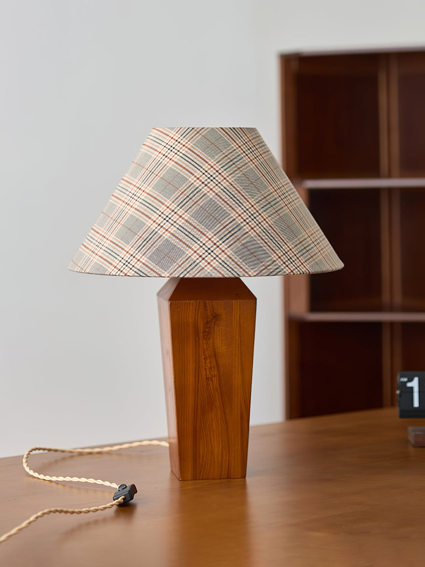 Plaid shade lamp unlit on a wood desk in front of a dark bookcase