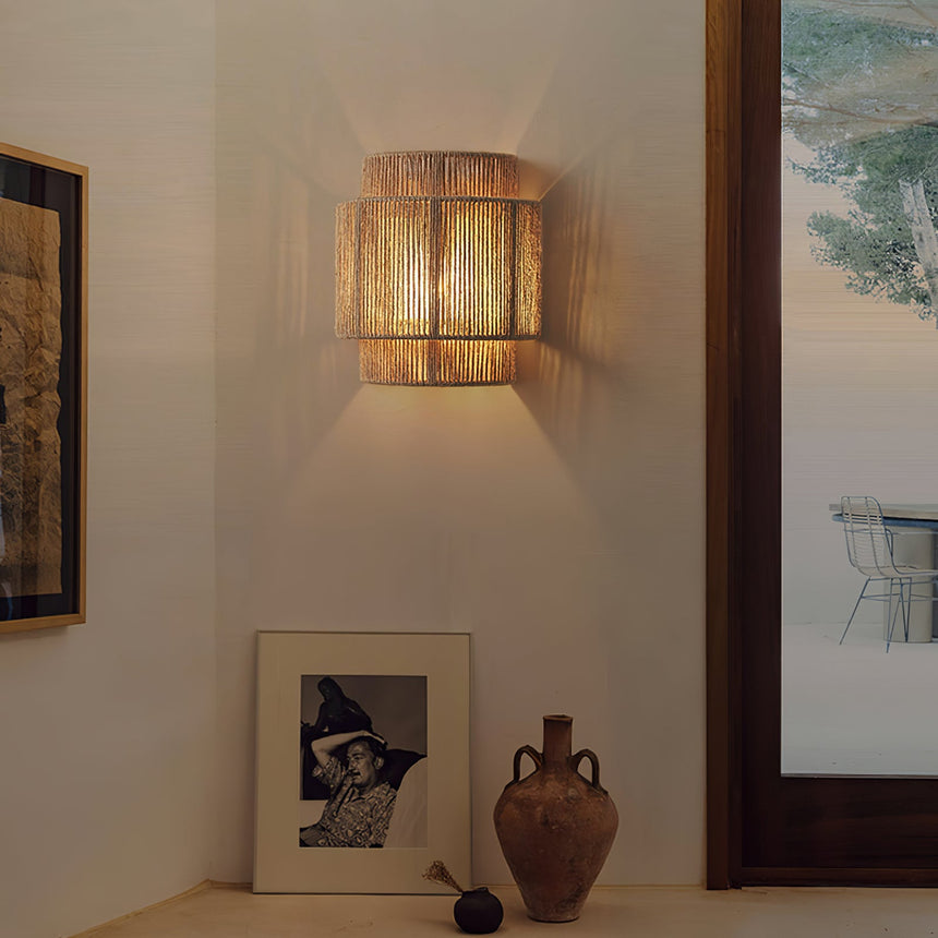 The hemp lamp lit on a white wall above a framed photo and a clay vase