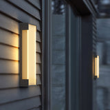 The sconce lit on gray lap siding beside a dark window reveal