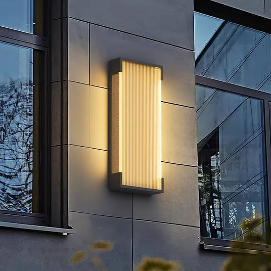 The sconce lit on a gray panel facade beside a tall window at dusk