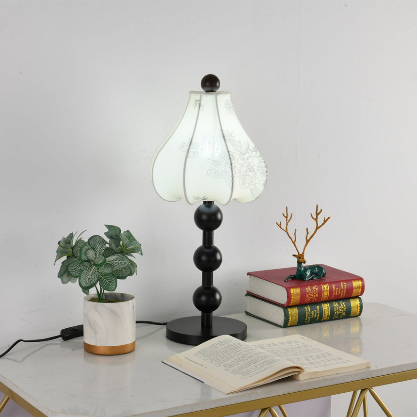 Floral table lamp unlit on a console beside stacked books and a plant