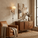 The lit lamp above a walnut cabinet beside a leather armchair and art