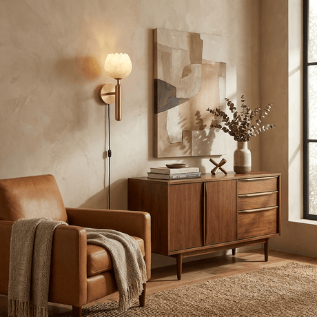 The lit lamp above a walnut cabinet beside a leather armchair and art