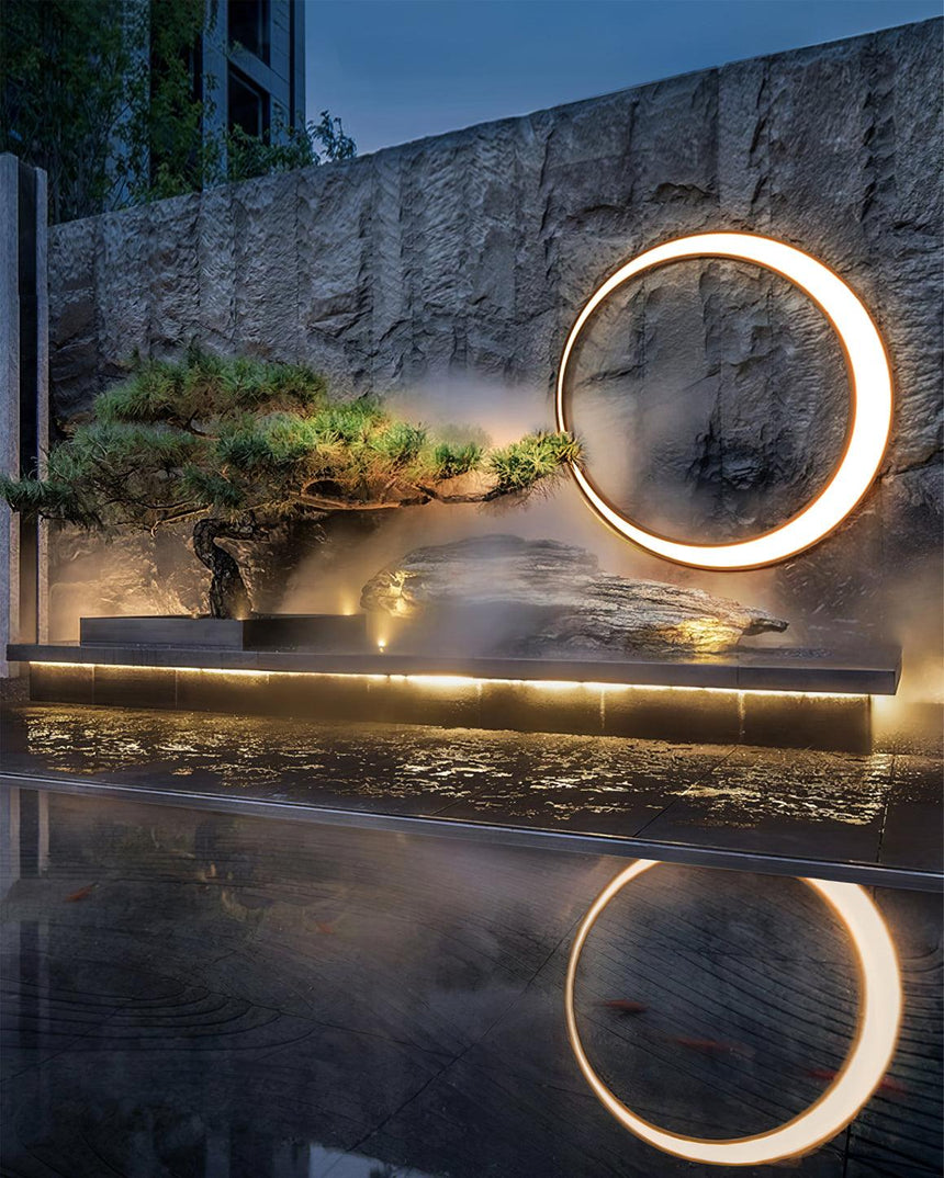 The ring lit on a textured wall above a reflecting pool and a pine