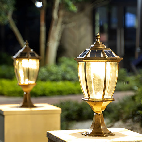 A bronze lantern lit on a pillar at dusk with shrubs behind it